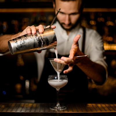 Professional bartender pouring a white cocktail from the steel shaker to the glass through the sieve on the bar counter in the blurred background