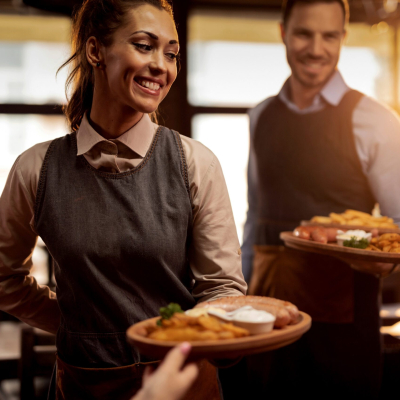 Two waiters serving lunch and brining food to their gusts in a tavern. Focus is on happy waitress.